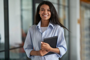 A young professional standing in an office hallway while holding a tablet.